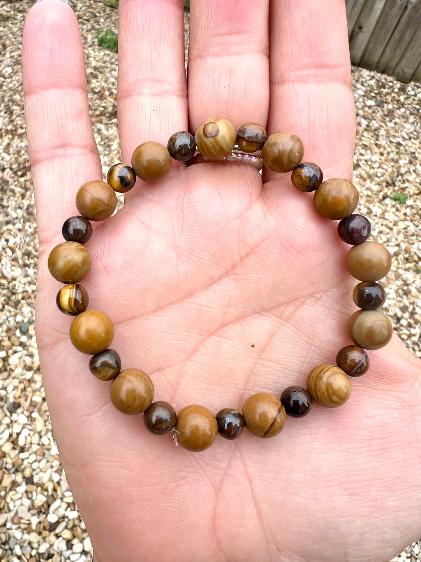 Hand wearing a beaded Wooden Jasper & tiger eye crystals bracelet with a pebbly surface behind