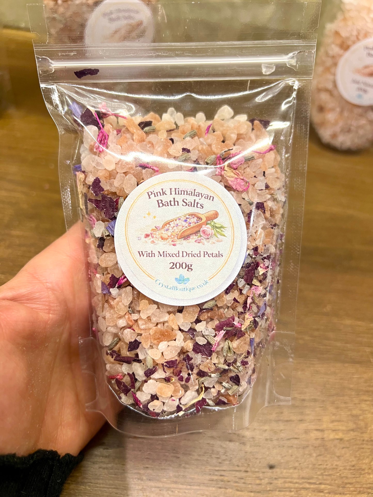 Packaged Pink Himalayan Bath Salts with Mixed Dried Petals held by a hand on a wooden surface.