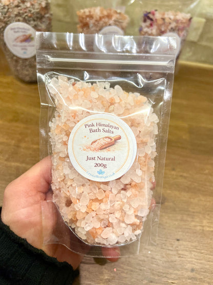 Hand holding a package of Pink Himalayan Bath Salts labeled 'Just Natural' on a wooden surface.