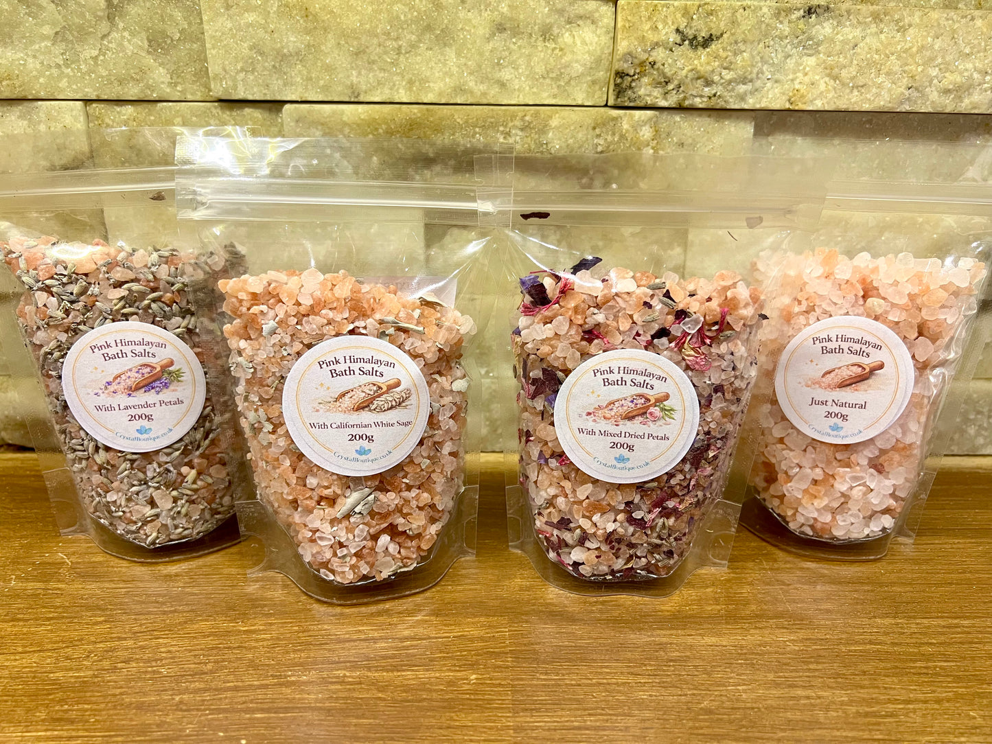 Four packages of pink Himalayan bath salts on a wooden surface with a stone wall background.