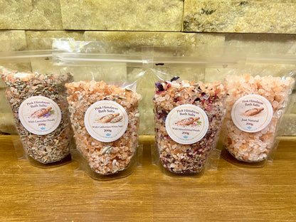 Four packages of pink Himalayan bath salts on a wooden surface with a stone wall background.