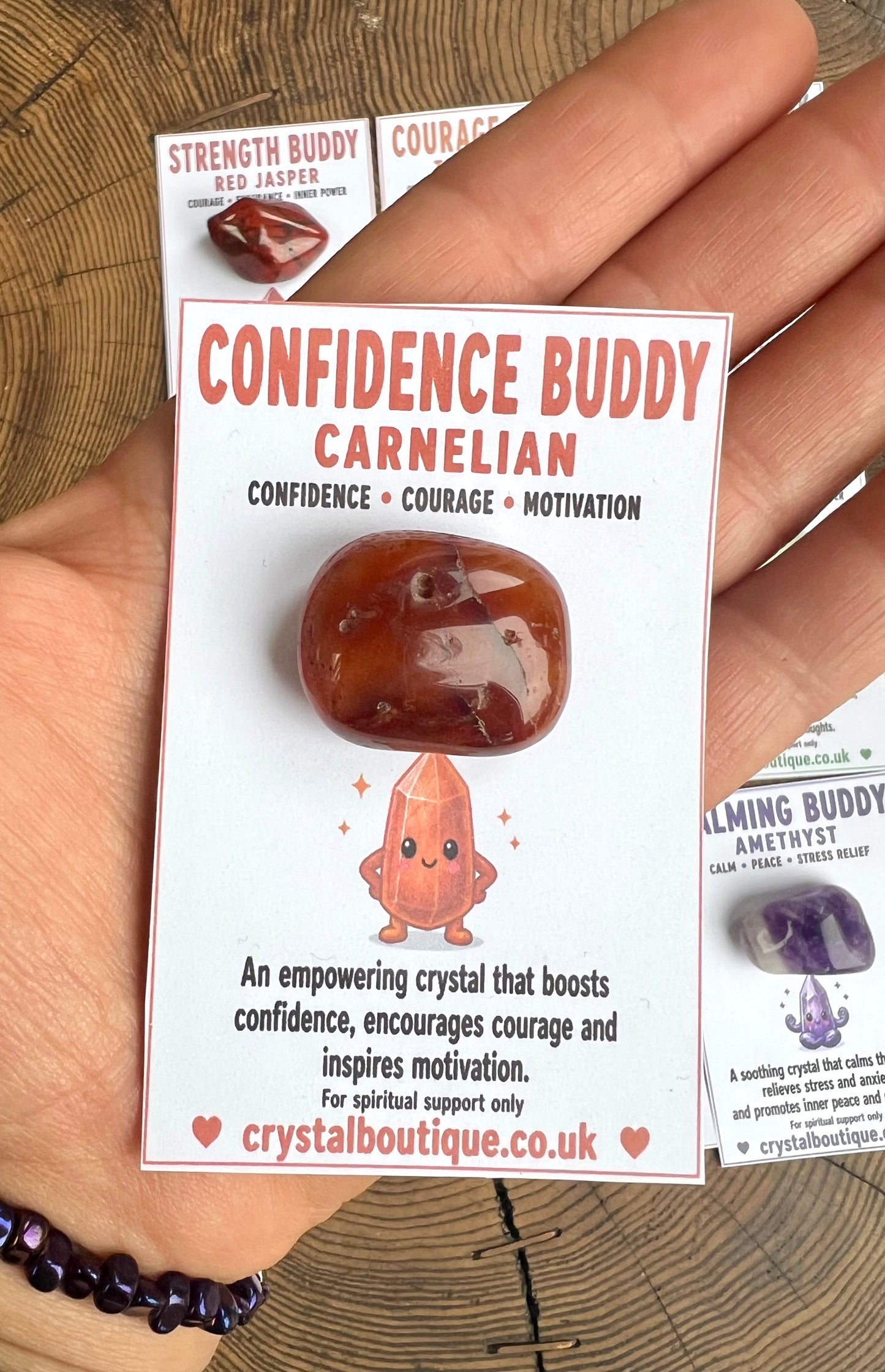 Carnelian crystal with packaging labeled 'Confidence Buddy' held by a hand on a wooden surface.