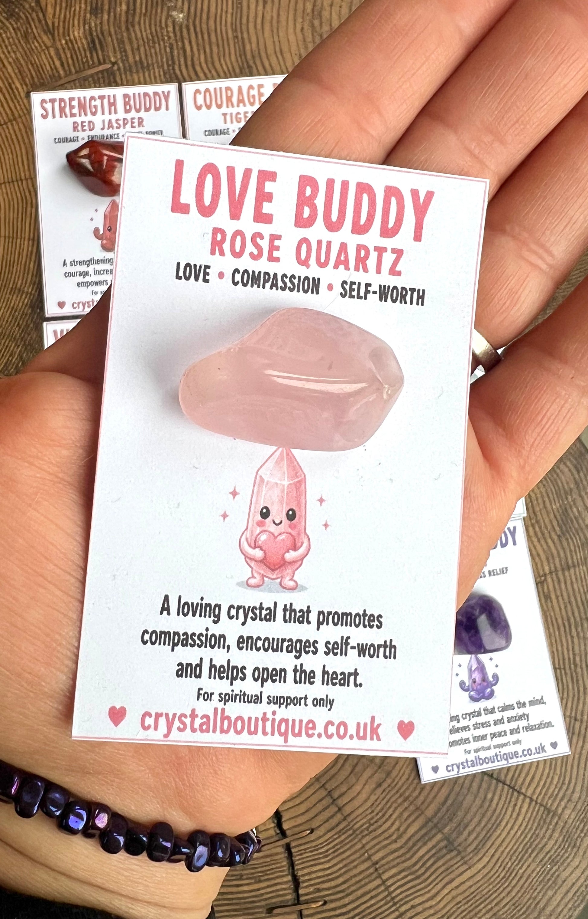 Love Buddy Rose Quartz crystal with packaging held by a hand on a wooden surface.