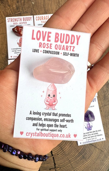 Love Buddy Rose Quartz crystal with packaging held by a hand on a wooden surface.