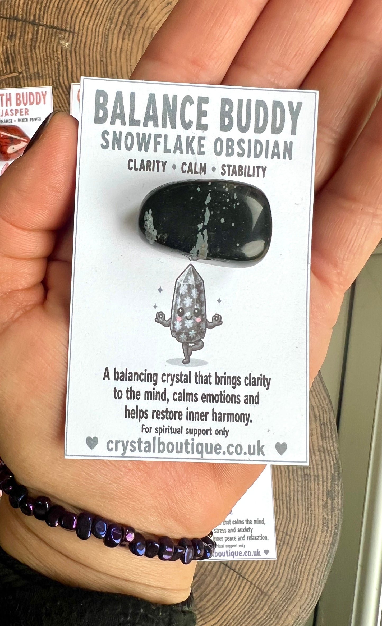 Hand holding a card with 'Balance Buddy Snowflake Obsidian' crystal information.