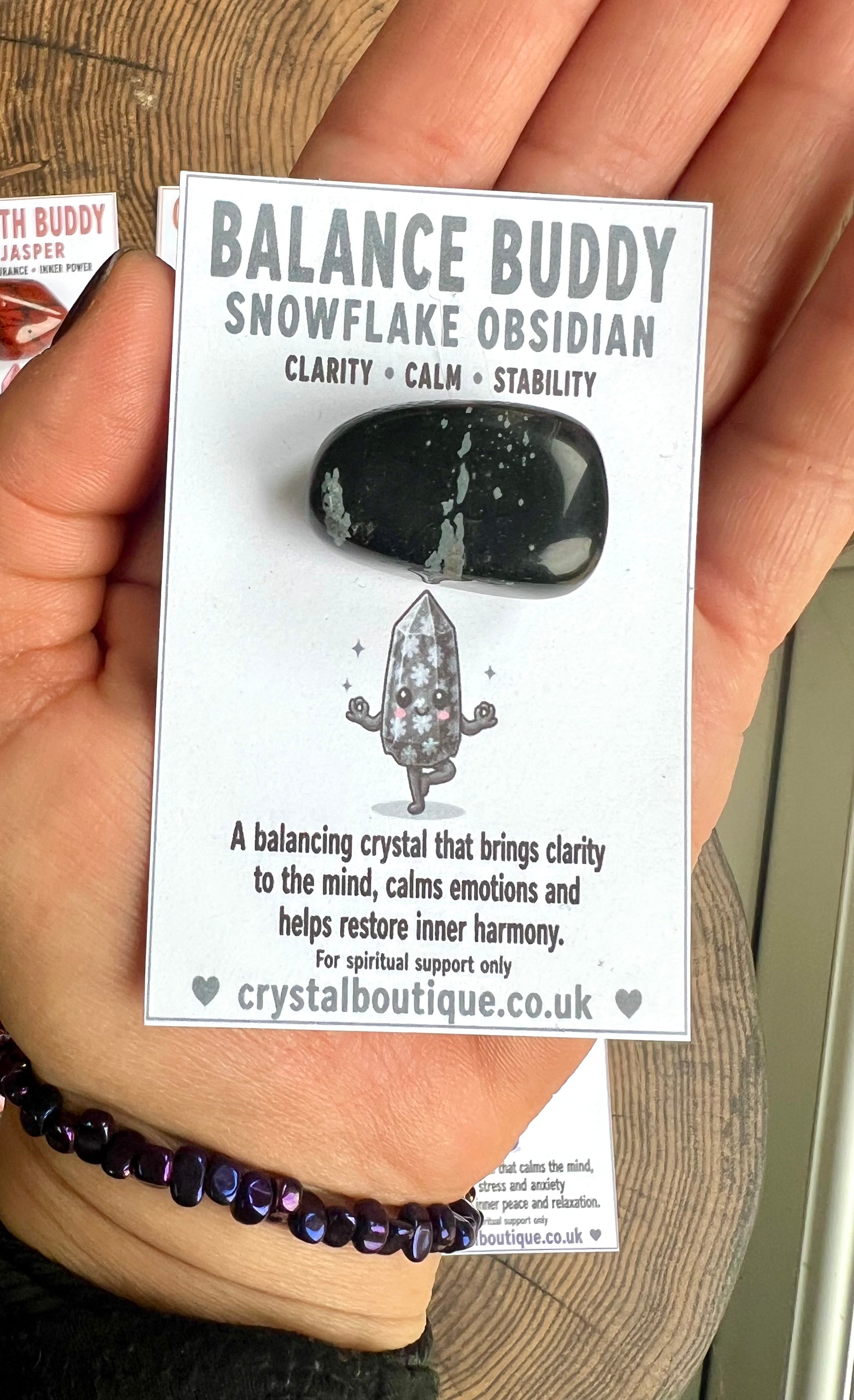 Hand holding a card with 'Balance Buddy Snowflake Obsidian' crystal information.