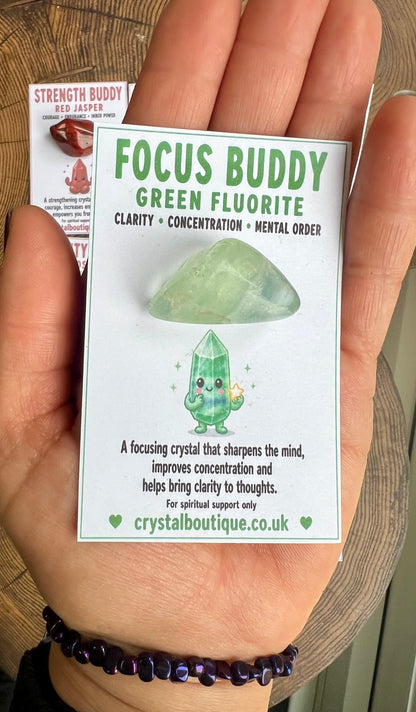Hand holding a 'Focus Buddy Green Fluorite' crystal packaging with text and a cartoon character on a wooden surface.