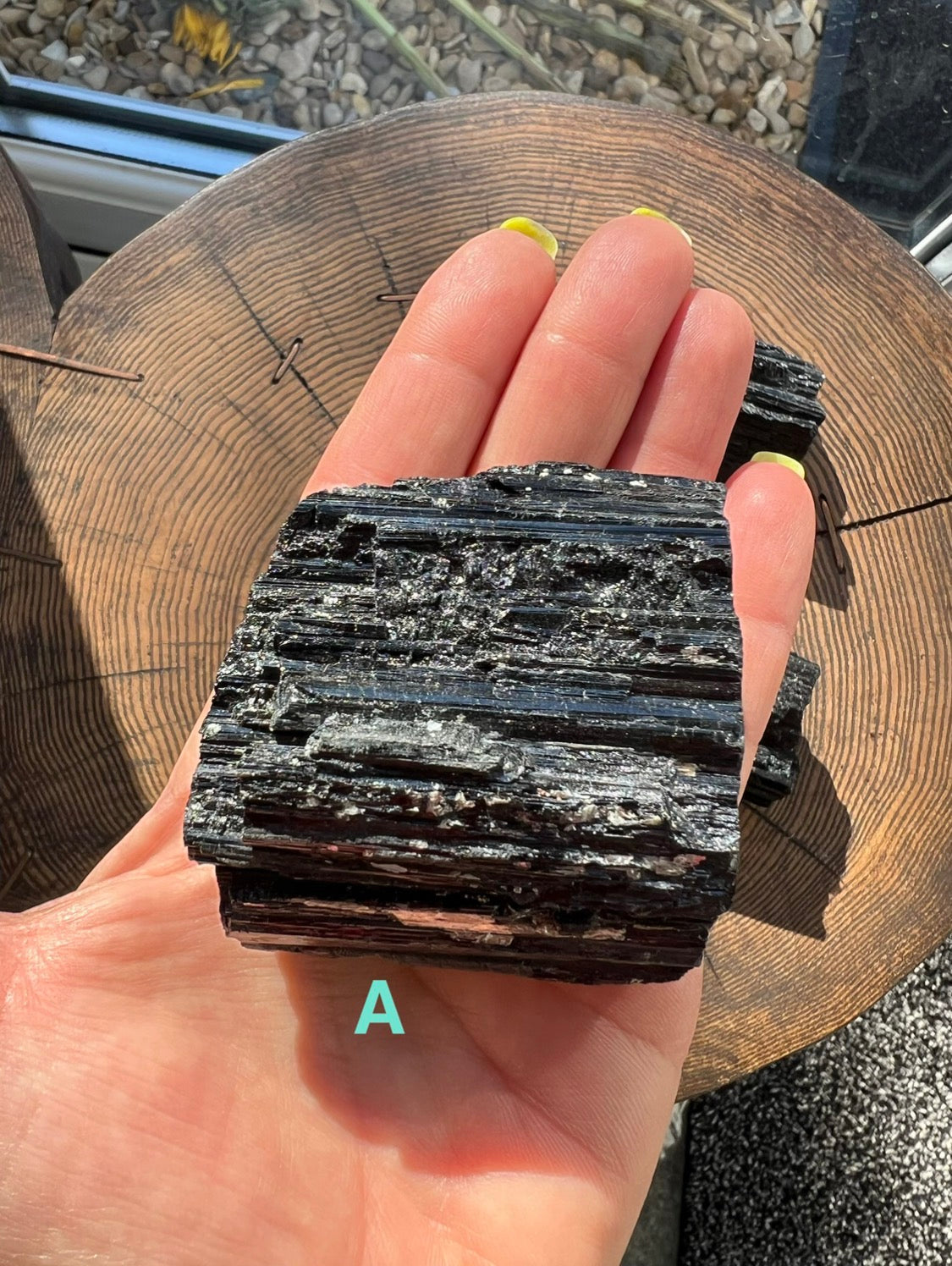 A hand holding a rough, dark black Tourmaline mineral with layered striated texture, photographed over a wooden surface. A turquoise letter 'A' appears near the bottom of the hand.