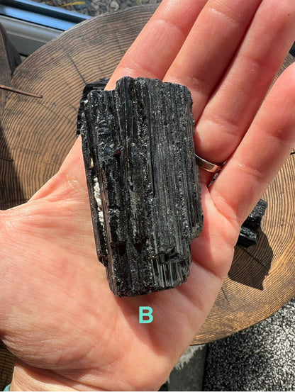 A hand holding a rough, dark black Tourmaline mineral with layered striated texture, photographed over a wooden surface. A turquoise letter 'B' appears near the bottom of the hand.