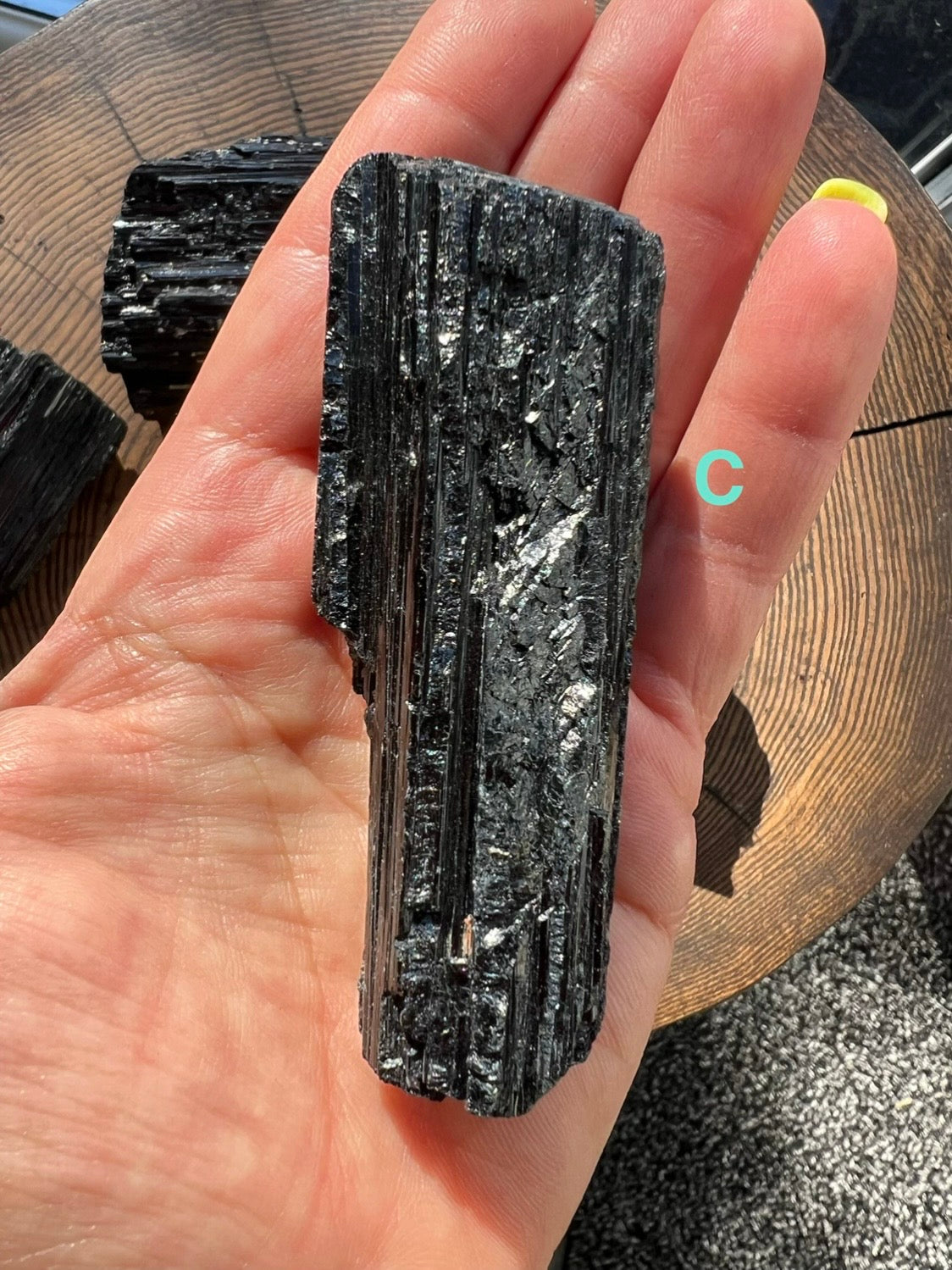 A hand holding a rough, dark black Tourmaline mineral with layered striated texture, photographed over a wooden surface. A turquoise letter 'c' appears near the side of the hand.