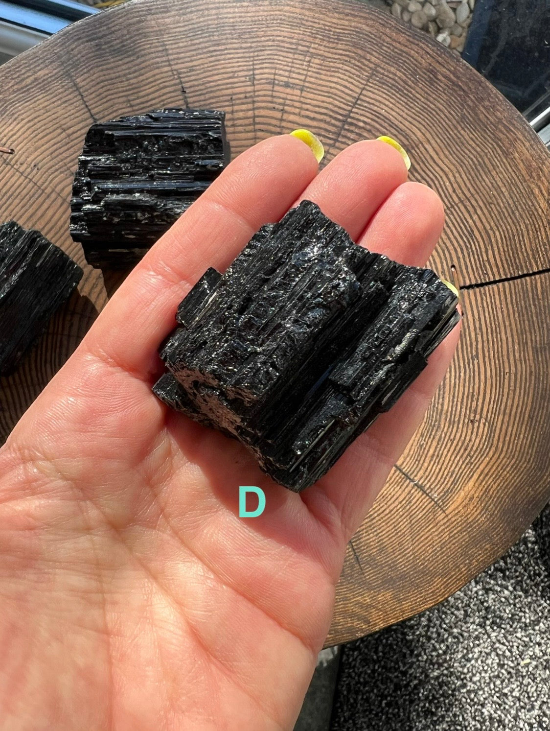 A hand holding a rough, dark black Tourmaline mineral with layered striated texture, photographed over a wooden surface. A turquoise letter 'D' appears near the bottom of the hand.