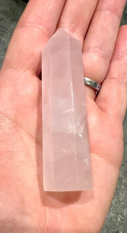 A pale pink rose quartz crystal point held in a person's hand, with a silver ring visible on one finger.