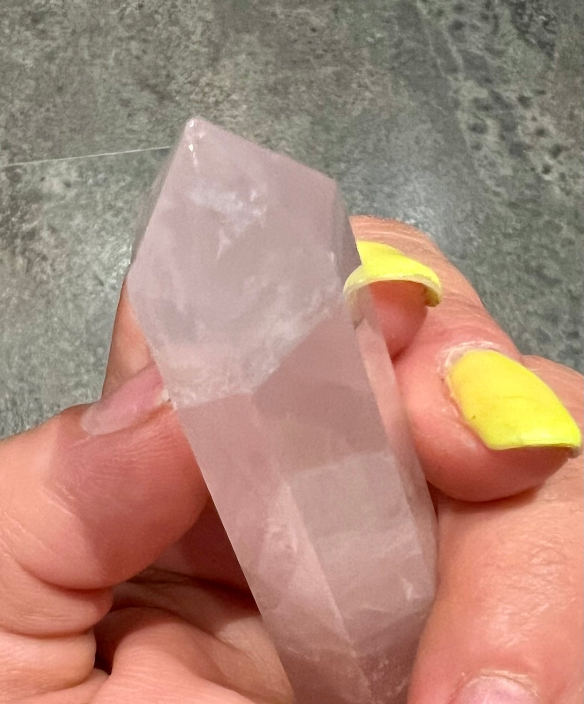 A hand holding a rose quartz crystal point against a gray stone tile background.