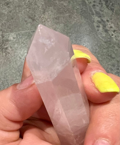 A hand holding a rose quartz crystal point against a gray stone tile background.