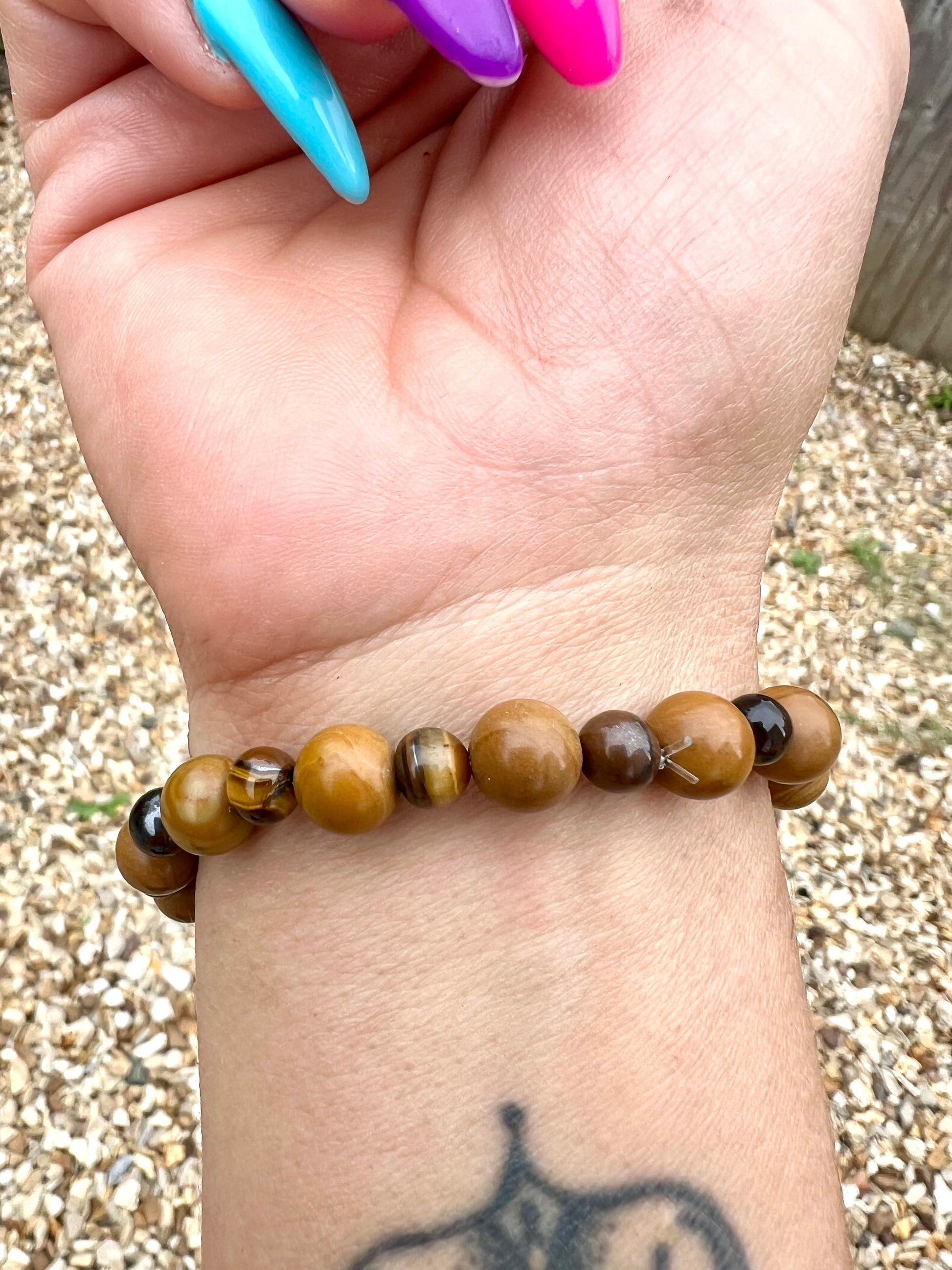 Hand wearing a beaded Wooden Jasper & tiger eye crystals bracelet with a pebbly surface behind