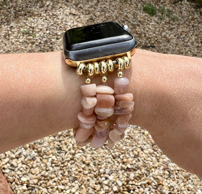 Peach Moonstone Crystal Apple watch strap with a gold band on a wrist, surrounded by pebbles.