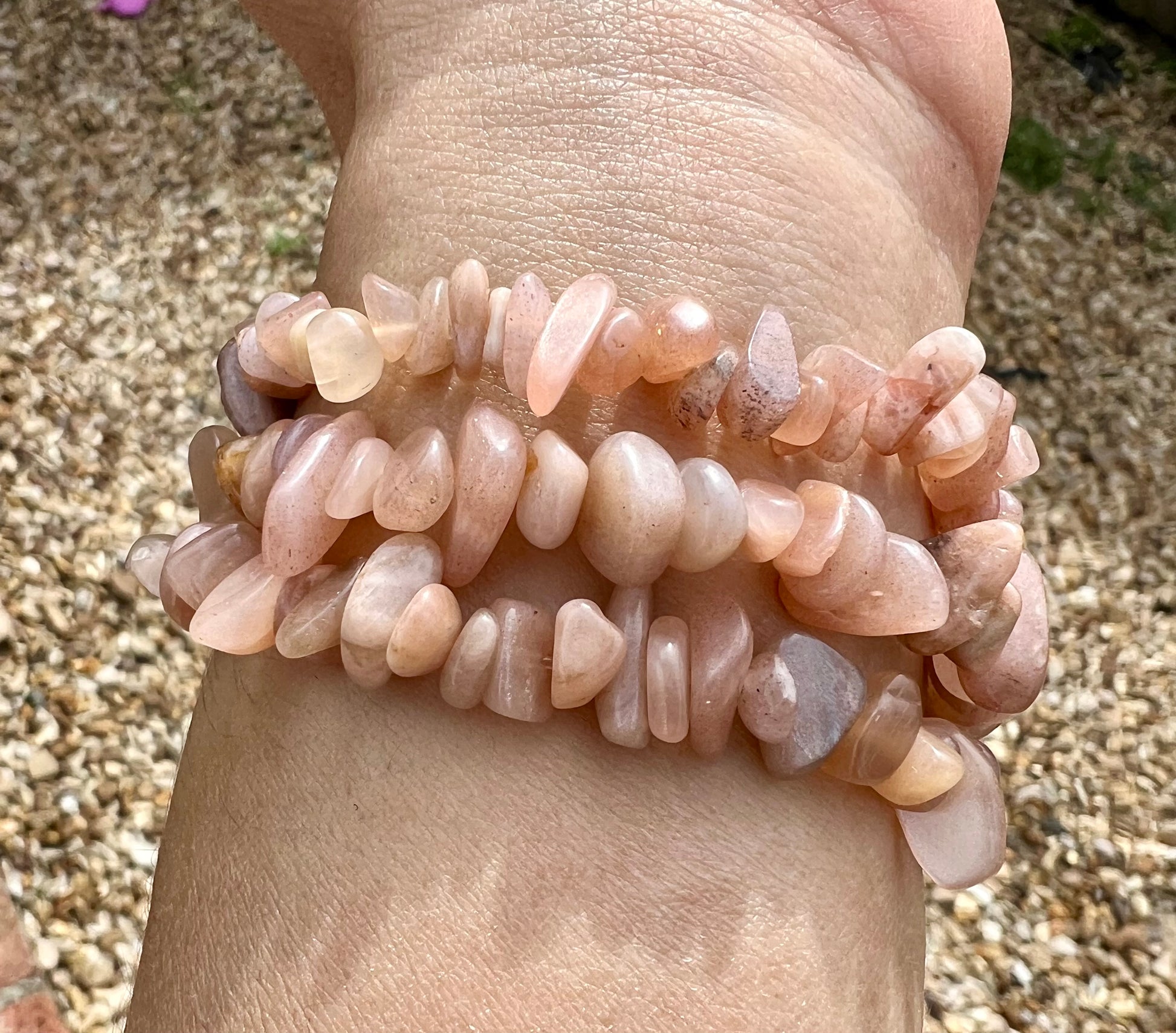 Peach Moonstone Crystal Apple watch strap with a gold band on a wrist, surrounded by pebbles.
