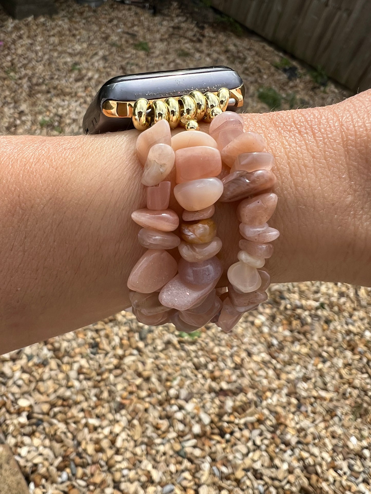 Peach Moonstone Crystal Apple watch strap with a gold band on a wrist, surrounded by pebbles.