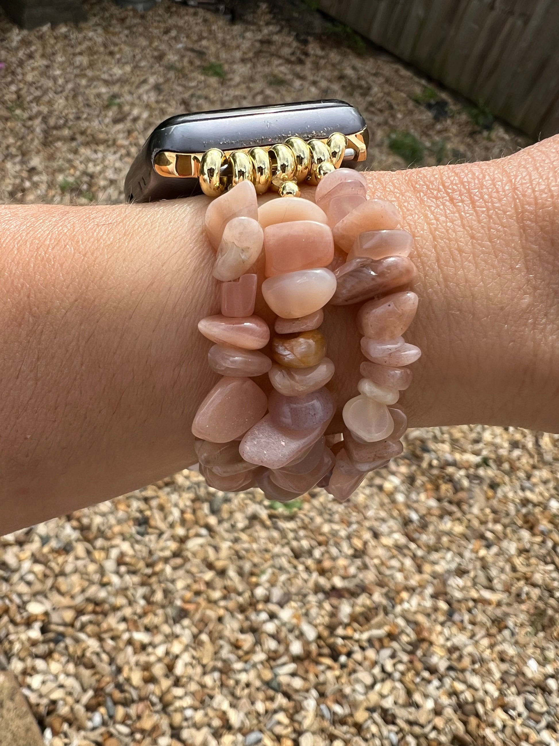 Peach Moonstone Crystal Apple watch strap with a gold band on a wrist, surrounded by pebbles.
