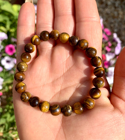 A hand holding a bracelet composed of 8mm round golden-brown Tiger Eye crystal gemstones.